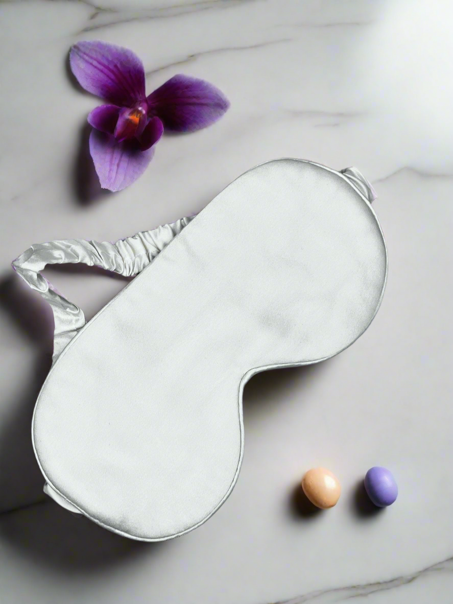 Antifaz para dormir de seda morera color blanco junto a una orquídea morada sobre mármol