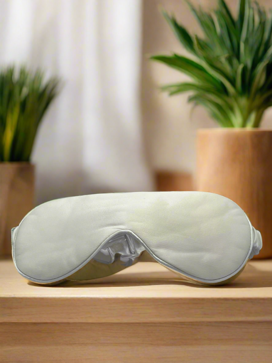 Antifaz para dormir de seda morera color blanco sobre una mesa de madera con plantas al fondo