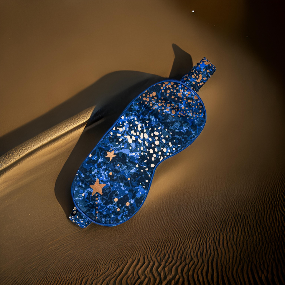 Antifaz de seda Cielo Nocturno en azul con estampado de estrellas doradas sobre un fondo de dunas.