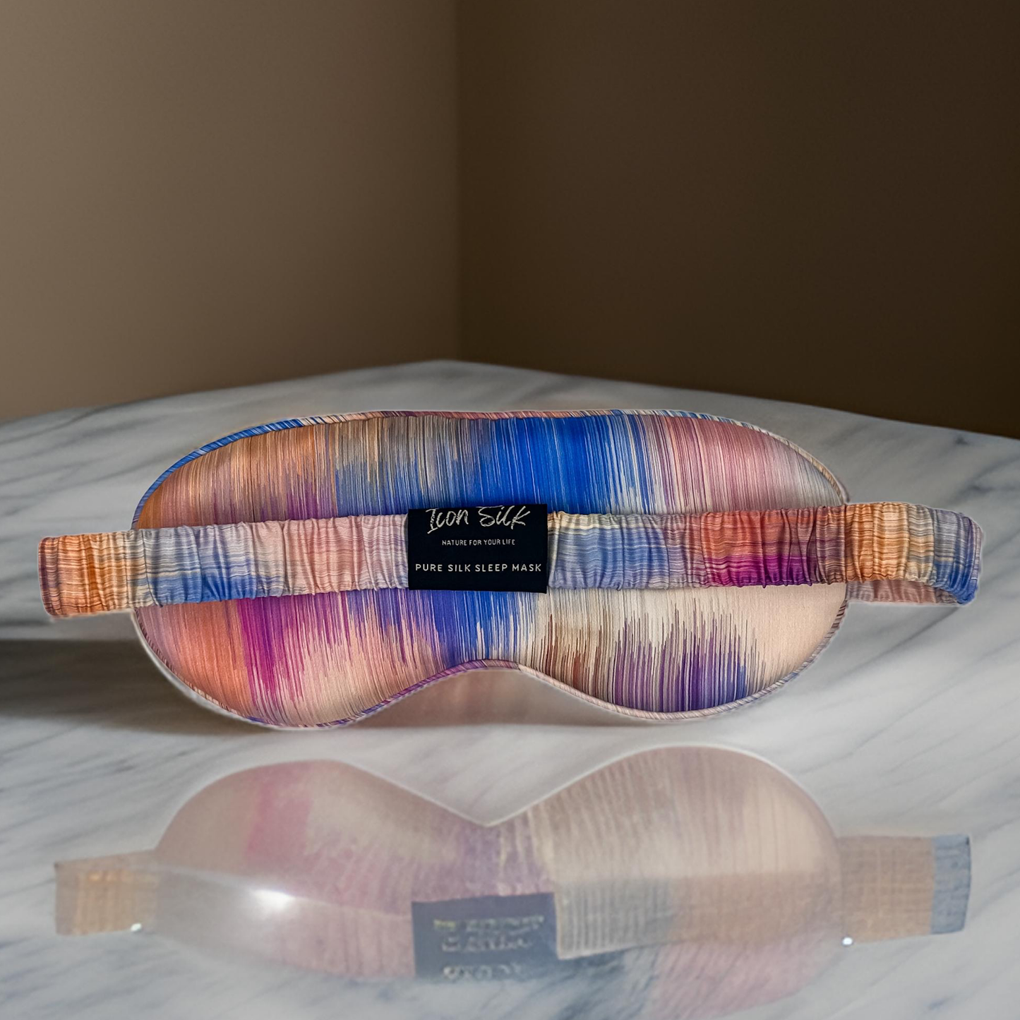 Parte posterior del antifaz para dormir de seda morera con estampado efecto amanecer multicolor, banda elástica y etiqueta Icon Silk sobre una superficie reflectante