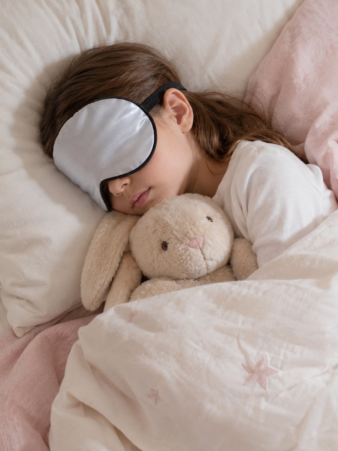 Niña durmiendo con un antifaz de seda gris y un peluche de conejo