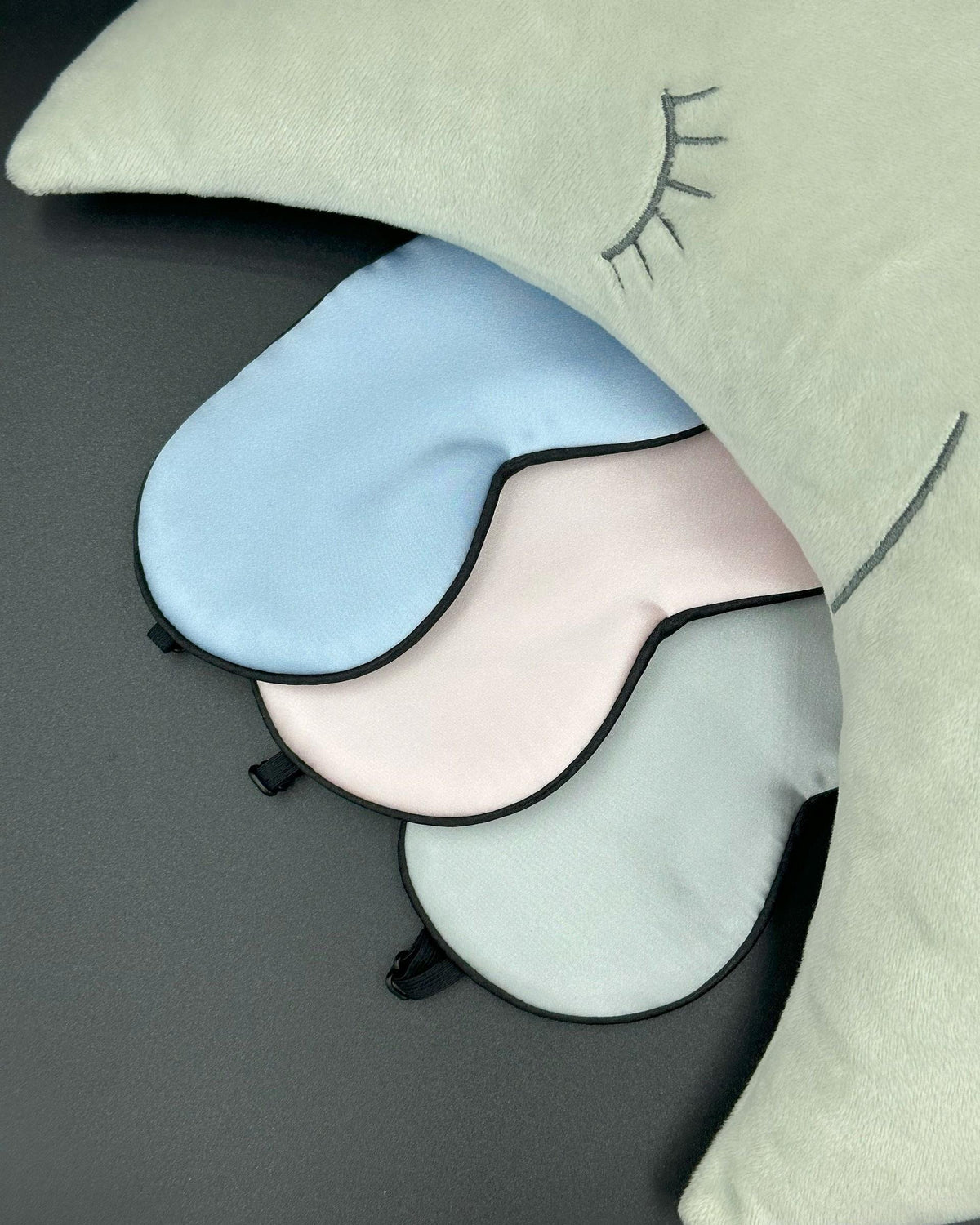 Antifaces de seda infantiles en azul claro, rosa y gris perla junto a un cojín en forma de luna