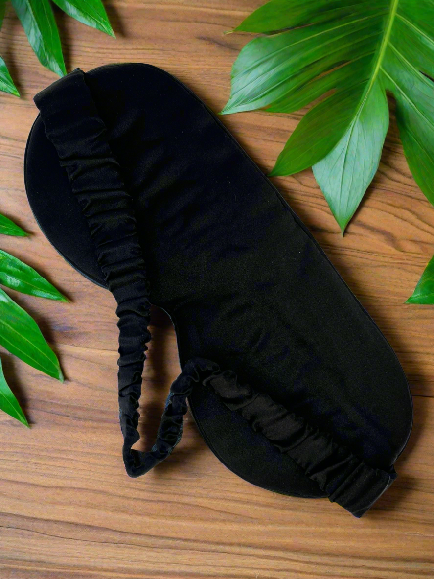 Antifaz para dormir de seda morera color negro con banda elástica sobre madera y rodeado de hojas verdes