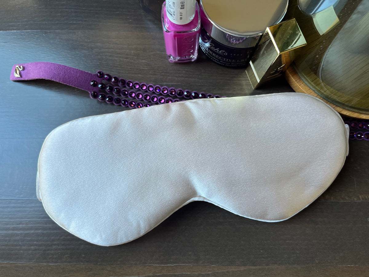 Antifaz para dormir de seda morera color nude sobre un tocador junto a un espejo y productos de belleza